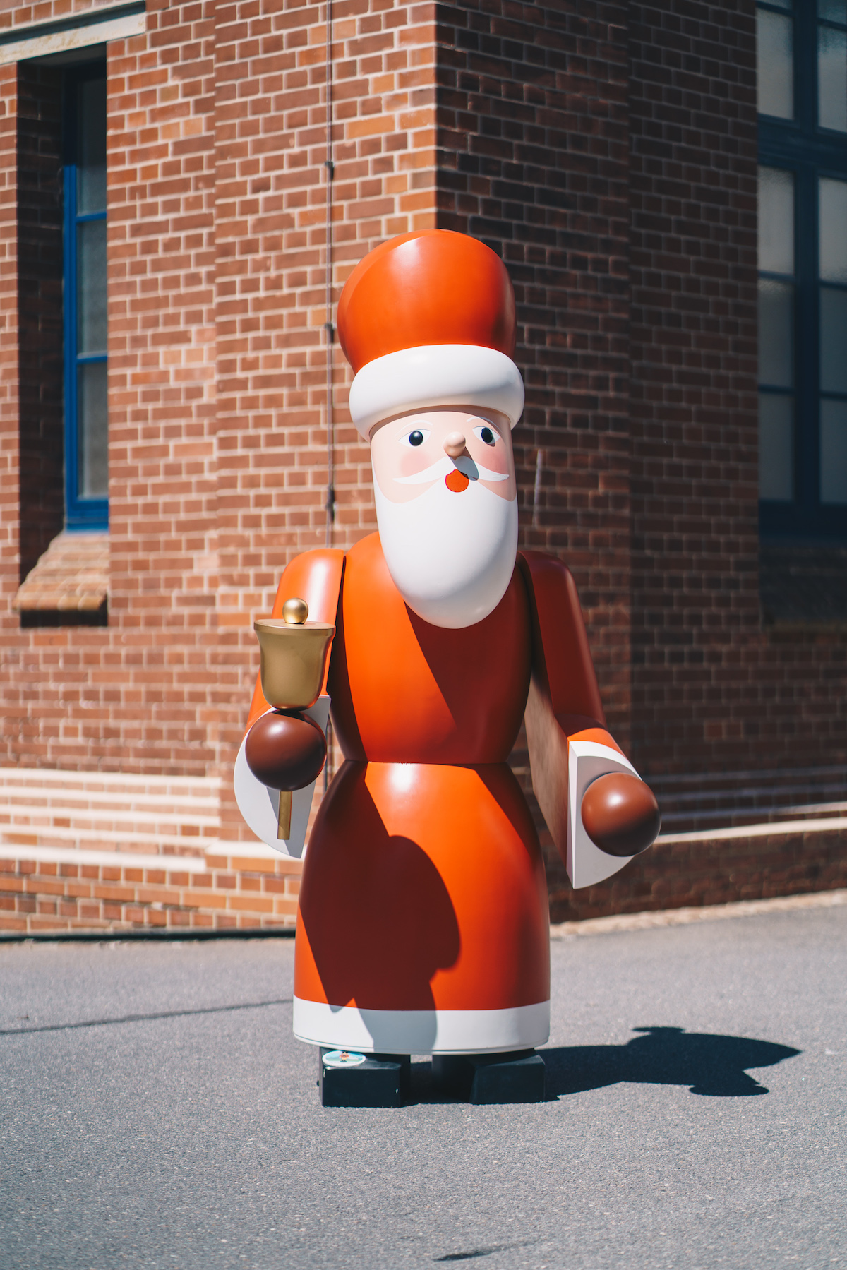 A red life-size Santa Claus made of wood stands in front of a wall of bricks.