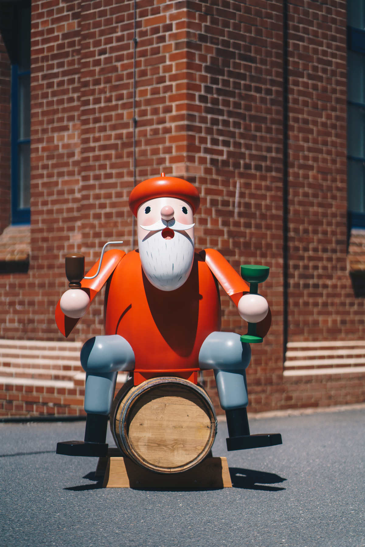 A red life-size wooden smoker sits on a wine barrel. This stands in front of a brick wall.